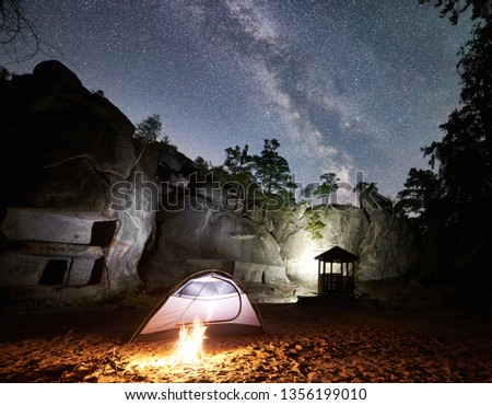 Tourist camping at summer night near mountain rocks. Glowing tent and campfire under magical night sky full of stars and Milky way. On the background beautiful starry sky, big boulders and trees