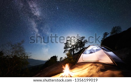 Camping at summer night on rocky mountain. White tourist tent and bonfire under magical night sky full of stars and Milky way. On the background beautiful starry sky, full moon, big boulders and trees