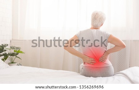 Senior woman suffering from backache in morning sitting on bed, red sore zone, panorama with free space Royalty-Free Stock Photo #1356269696