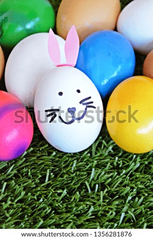 A white egg with a rabbit face painted on it and ears glued on it lies with other different eggs on a grass field - concept for Easter with place for text