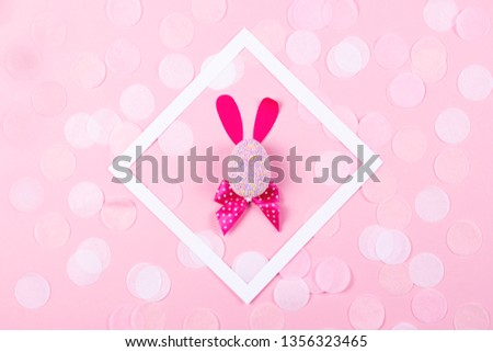 Easter bunny made of an egg with paper ears and dotted magenta bow inside white square frame decorated with festive confetti on pastel pink background. Easter holiday and tradition concept. Flat lay.