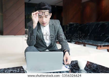 Wow! Surprised young asian businessman with opened mouth touching glasses, glad and shocked about his work while looking at his computer. Sitting at the lobby area of a modern hotel.Copy space.
