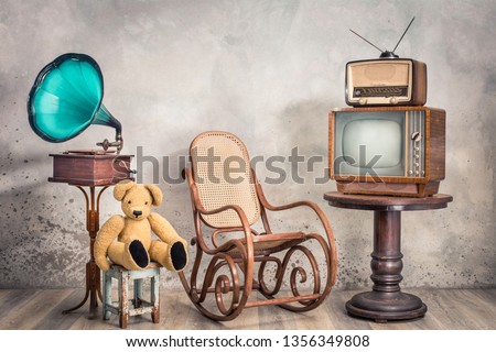 Retro TV receiver and old broadcast radio from circa 50s on wooden table, outdated gramophone, Teddy Bear toy, aged rocking chair front textured concrete wall background. Vintage style filtered photo