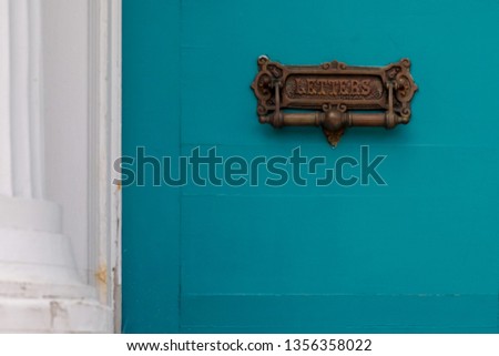 A vintage brass letters mail slot and door knocker. The door is a teal color with a white pillar and door frame. The old worn decorative metal plate has the word letters on the mail slot of a house. 