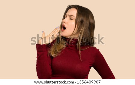 Teenager girl with turtleneck yawning and covering wide open mouth with hand over isolated ocher background