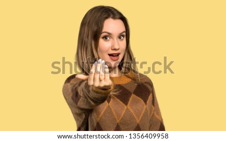 Teenager girl with brown sweater inviting to come with hand. Happy that you came over isolated yellow background
