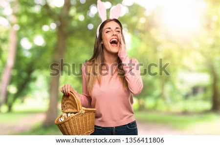 Young woman wearing bunny ears for Easter holidays shouting with mouth wide open and announcing something in the park