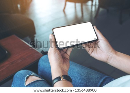 Mockup image of woman's hands holding black mobile phone with blank white screen horizontally in cafe