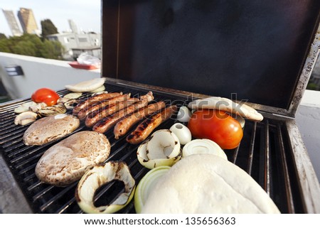 Sausages, onion slices, tomatoes and pita bread getting ready on an outdoor rooftop barbecue grill. Some view of the city blirred in the background.