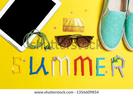 blue slip on shoes, earphones ,sunglasses ,credit card and a smartphone. word summer made from constructor parts is layed near on a yellow surface. Top view