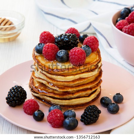 Homemade pancakes with berries and honey on a pink plate over white wooden surface, side view. Close-up. 