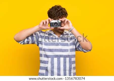 Blonde man over yellow wall holding a camera