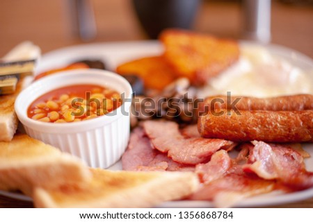 Full English Breakfast Close up with Shallow Focus with Fried Eggs, Baked Beans, Bacon, Mushrooms, Hash Browns, Sausages, Tomato, Black Pudding, Toast and Butter Portions