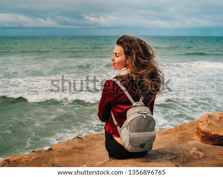 A girl in a red sweatshirt stands and her long hair flutters in the wind on a rock in San Diego overlooking the ocean line
