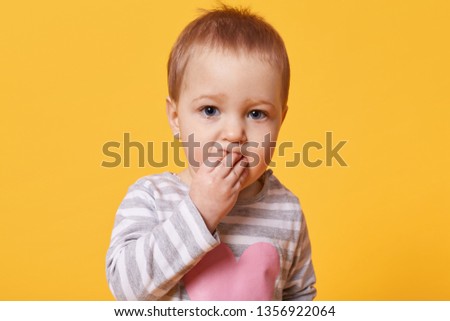 Portrait of a cute brooding girl with short fair hair keeping fingers in her mouth. A serious kid stands in front of camera looking straight, feels bored, search for new funny pastime. Family concept.