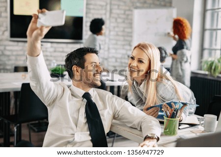 Two happy business people using smart phone and taking selfie while working in the office. There are people in the background. 