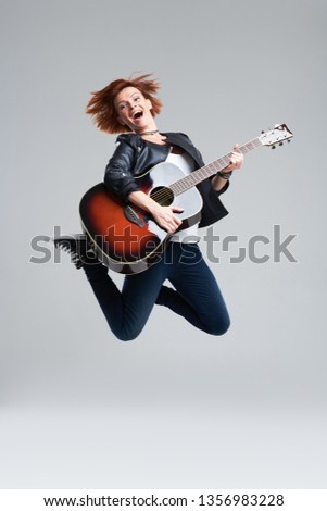 Young woman musician with an acoustic guitar in hand on a gray background. She laughs and jumps high. plays rock and roll loudly. Full-length portrait.