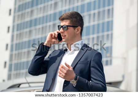 Young businessman wearing sunglasses standing near car answering phone call from business partner explaining situation side view