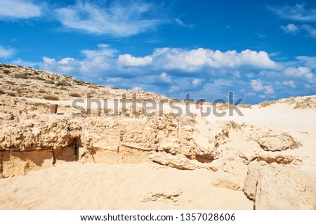 Image of one young white girl standing on a rough limestone cliff near Cape Greco, Cyprus. Many white clouds in the sky, barren orange sandy hill, vast expanse. Warm day in fall