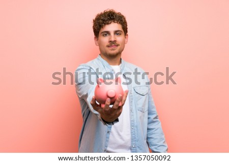 Blonde man over pink wall holding a piggybank