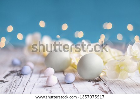 Natural colored Easter eggs, malt candy covered chocolate eggs, and flower blossoms over a rustic white wood table against a blue background.