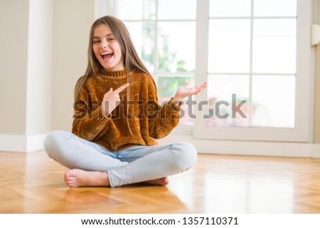 Beautiful young girl kid sitting on the floor at home amazed and smiling to the camera while presenting with hand and pointing with finger.