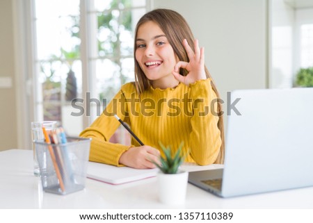 Beautiful young girl studying using computer laptop and writing on notebook doing ok sign with fingers, excellent symbol
