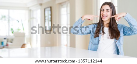 Wide angle picture of beautiful young woman sitting on white table at home smiling confident showing and pointing with fingers teeth and mouth. Health concept.