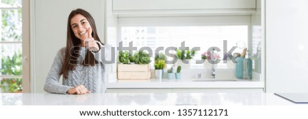 Wide angle picture of beautiful young woman sitting on white table at home doing happy thumbs up gesture with hand. Approving expression looking at the camera with showing success.