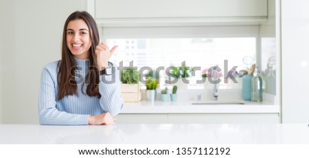 Wide angle picture of beautiful young woman sitting on white table at home smiling with happy face looking and pointing to the side with thumb up.