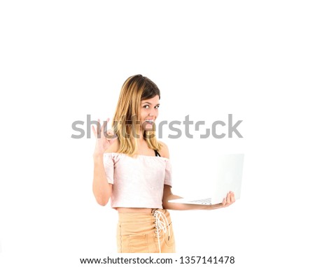 Happy young blonde teenager girl doing an ok gesture with her hand while holding a laptop against a white background