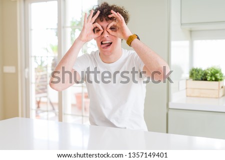 Young handsome man wearing white t-shirt at home doing ok gesture like binoculars sticking tongue out, eyes looking through fingers. Crazy expression.