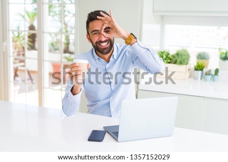 Handsome hispanic man working using computer laptop and drinking a cup of coffee with happy face smiling doing ok sign with hand on eye looking through fingers
