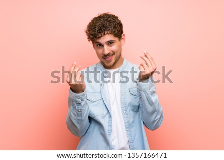 Blonde man over pink wall making money gesture