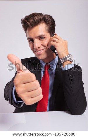 young business man sitting at the desk and talking on the phone while showing thumbs up with a smile on his face