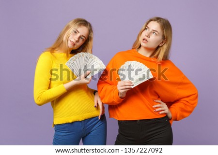 Two pensive blonde twins sisters girls in colorful clothes holding fan of money in dollar banknotes, cash money isolated on violet blue background. People family lifestyle concept. Mock up copy space