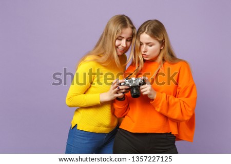 Two pretty young blonde twins sisters girls in vivid colorful clothes holding retro vintage photo camera isolated on violet blue wall background. People family lifestyle concept. Mock up copy space
