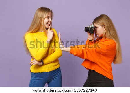 Two young blonde twins sisters girls in vivid colorful clothes taking pictures on retro vintage photo camera isolated on violet blue background. People family lifestyle concept. Mock up copy space