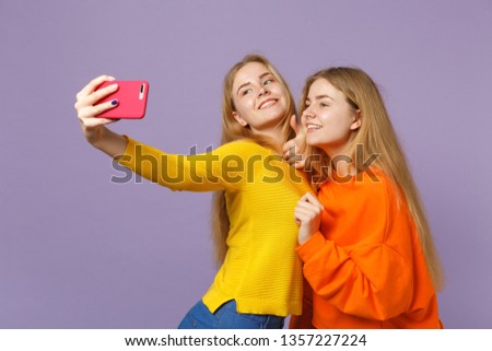 Two smiling young blonde twins sisters girls in colorful clothes doing selfie shot on mobile phone isolated on pastel violet blue wall background. People family lifestyle concept. Mock up copy space