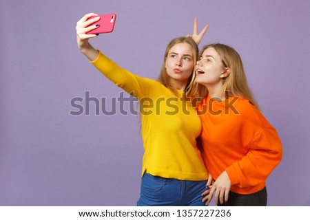 Two cheerful young blonde twins sisters girls in colorful clothes doing selfie shot on mobile phone isolated on pastel violet blue wall background. People family lifestyle concept. Mock up copy space