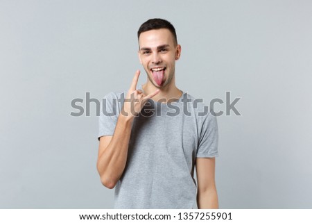 Funny young man in casual clothes showing tongue, depicting heavy metal rock sign gesture isolated on grey background. People sincere emotions, lifestyle concept. Mock up copy space. Advertising area