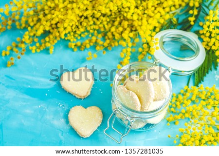 Delicious baked Heart shaped cookies with coconut chips in glass jar and fresh silver wattle or mimosa on blue concrete table surface. Food greeting card with copy space for you text.