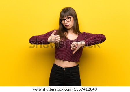 Woman with glasses over yellow wall making good-bad sign. Undecided between yes or not