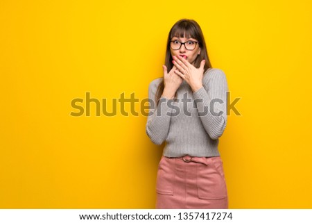 Woman with glasses over yellow wall covering mouth with hands for saying something inappropriate