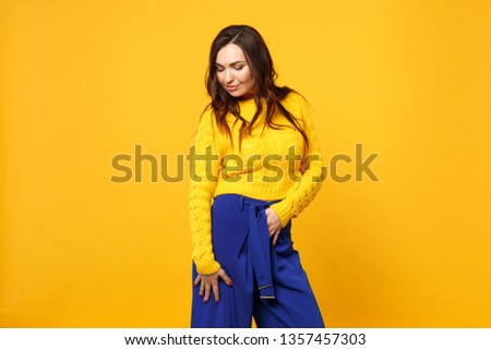 Portrait of tender young woman in sweater, blue trousers standing and looking down isolated on yellow orange wall background in studio. People sincere emotions, lifestyle concept. Mock up copy space