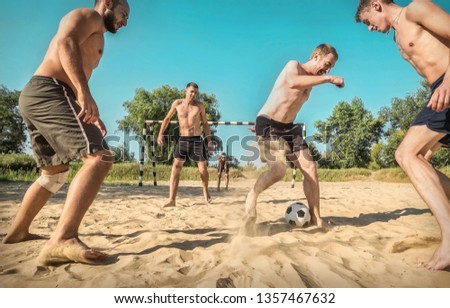 Happy young people in active dynamic action with ball funny play in football on the beach under sun light in summer time and in sunny day. Soccer on the sand.