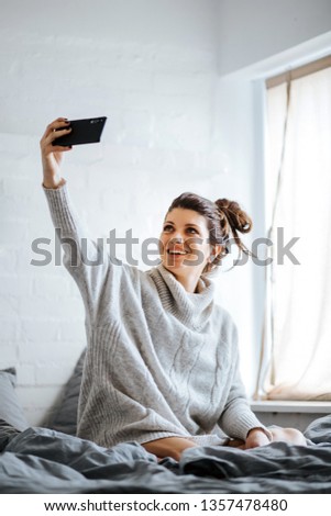 Portrait of a young, beautiful brunette taking selfies in her bed next to the window