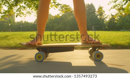 CLOSE UP, SUN FLARE: Unrecognizable young woman skating in the green park on her electric skateboard. Cinematic close up shot of a sporty girl cruising on an e-longboard on beautiful summer afternoon