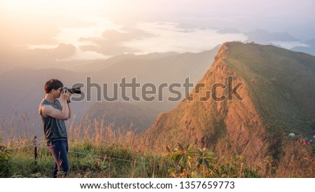 Young Asian male photographer and traveler taking photo of mountain landscape scenery during sunrise at Khao San Nok Wua in Kanchanaburi, Thailand. Travel photography concept