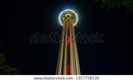 Upward view of the Tower of the Americas at night with red navigational lights with blue/green/white accent lights around the bottom/middle of the observation deck. Some foliage in the foreground.